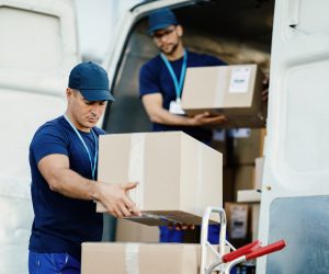 Young courier and his colleague unloading cardboard boxes from delivery van.